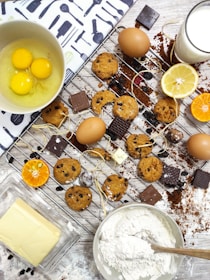 A baking scene featuring cookies with chocolate chips on a cooling rack surrounded by whole eggs, chocolate pieces, lemon slices, a bowl of flour, butter, and a glass of milk. The setup includes a mix of ingredients and baked goods on a wooden surface with some spilled cocoa and flour for a rustic touch.