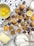 A baking scene featuring cookies with chocolate chips on a cooling rack surrounded by whole eggs, chocolate pieces, lemon slices, a bowl of flour, butter, and a glass of milk. The setup includes a mix of ingredients and baked goods on a wooden surface with some spilled cocoa and flour for a rustic touch.