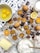 A baking scene featuring cookies with chocolate chips on a cooling rack surrounded by whole eggs, chocolate pieces, lemon slices, a bowl of flour, butter, and a glass of milk. The setup includes a mix of ingredients and baked goods on a wooden surface with some spilled cocoa and flour for a rustic touch.