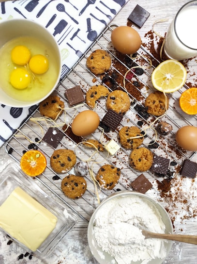 A baking scene featuring cookies with chocolate chips on a cooling rack surrounded by whole eggs, chocolate pieces, lemon slices, a bowl of flour, butter, and a glass of milk. The setup includes a mix of ingredients and baked goods on a wooden surface with some spilled cocoa and flour for a rustic touch.