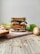 A tall sandwich stacked with multiple layers of ingredients such as lettuce, tomato slices, boiled egg slices, and various meats, placed on a wooden cutting board. Surrounding the sandwich are slices of bread, a tomato half, lettuce leaves, a potato, and scattered oats. The background is plain and minimalistic.