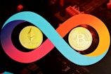 Two cryptocurrency coins, Ethereum and Bitcoin, are prominently displayed within a colorful infinity loop design. The Ethereum coin is positioned on the left, and the Bitcoin coin is on the right, both appearing gold and metallic against the vibrant swirling background of blue, red, and orange.