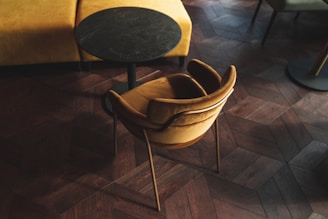 A cozy interior featuring a plush mustard-colored chair with an elegant design, positioned next to a small, round, black marble table. The setting rests on a dark, textured, herringbone-patterned wooden floor, adding sophistication to the ambiance.