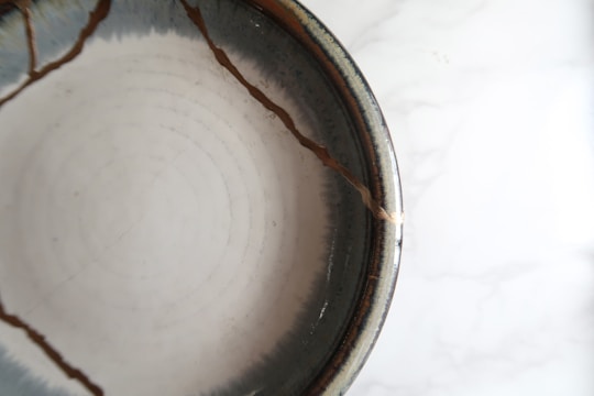A ceramic bowl with visible cracks repaired using a golden substance, likely representing the art of kintsugi. The bowl has a smooth, glossy texture with concentric circular patterns inside.