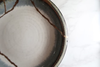 A ceramic bowl with visible cracks repaired using a golden substance, likely representing the art of kintsugi. The bowl has a smooth, glossy texture with concentric circular patterns inside.