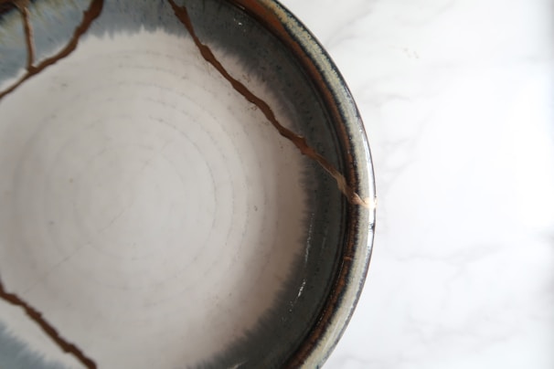 A ceramic bowl with visible cracks repaired using a golden substance, likely representing the art of kintsugi. The bowl has a smooth, glossy texture with concentric circular patterns inside.
