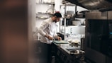 A chef is focused on preparing food in a commercial kitchen. He is wearing a white chef's coat and standing at a stainless steel counter, chopping ingredients on a green cutting board. The kitchen is equipped with various utensils, plates, and cooking appliances. There are shelves with plates and glasses in the background.