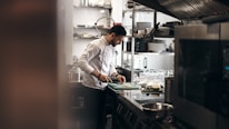 A chef is focused on preparing food in a commercial kitchen. He is wearing a white chef's coat and standing at a stainless steel counter, chopping ingredients on a green cutting board. The kitchen is equipped with various utensils, plates, and cooking appliances. There are shelves with plates and glasses in the background.