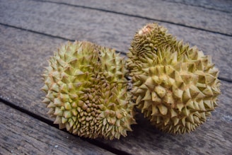 Two durian fruits with a spiky rind are resting side by side on a wooden surface. The surface appears weathered, adding texture to the background.