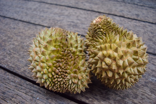 Two durian fruits with a spiky rind are resting side by side on a wooden surface. The surface appears weathered, adding texture to the background.