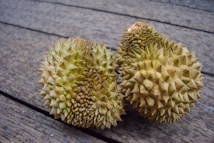 Two durian fruits with a spiky rind are resting side by side on a wooden surface. The surface appears weathered, adding texture to the background.