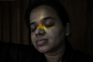 A person is resting with their eyes closed, experiencing a serene and peaceful moment. A spectrum of colorful light, resembling a rainbow, is cast across their face, creating an artistic and contemplative atmosphere. The background is dimly lit, emphasizing the contrast between the darkness and the vibrant colors on the person's face.