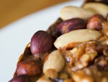 A close-up view of various types of nuts embedded in a baked item. The image features whole almonds and hazelnuts with a glossy, roasted appearance set against a textured brown surface.