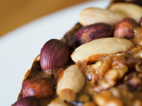 A close-up view of various types of nuts embedded in a baked item. The image features whole almonds and hazelnuts with a glossy, roasted appearance set against a textured brown surface.