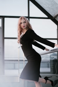 A woman with long blonde hair is wearing a fitted black dress and high heels. She is posed in a modern, minimalistic interior with large windows, creating a striking and elegant look.