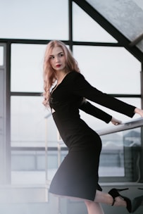 A sleek vertical photo of a woman wearing a minimalist black dress in a bright, white studio.