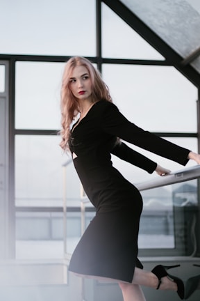 A woman with long blonde hair is wearing a fitted black dress and high heels. She is posed in a modern, minimalistic interior with large windows, creating a striking and elegant look.