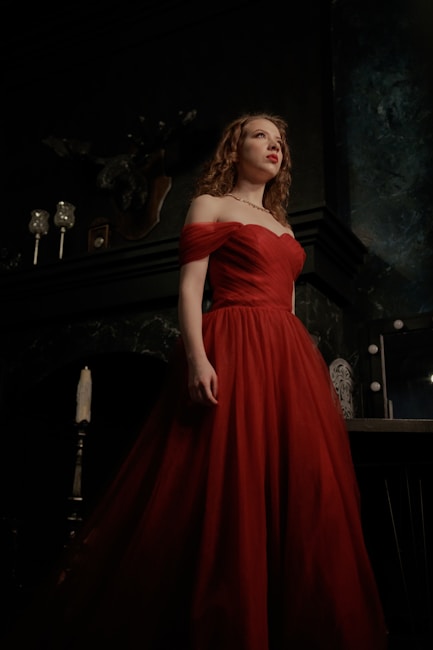 A woman in a flowing red gown stands confidently against a dark, elegant background. The setting appears to be lavish, with elements such as candles and ornate decor visible. Her expression is thoughtful and slightly upward gazing.