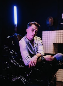 A person is seated in a dark room, illuminated by a bright, vertical blue light on the left side. The person is wearing a light-colored shirt and appears to be relaxed, leaning back slightly with one arm resting on the armrest. The room features a tiled wall with a subtle geometric pattern, adding to the modern setting.