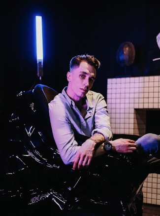 A person is seated in a dark room, illuminated by a bright, vertical blue light on the left side. The person is wearing a light-colored shirt and appears to be relaxed, leaning back slightly with one arm resting on the armrest. The room features a tiled wall with a subtle geometric pattern, adding to the modern setting.