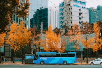 A bright blue bus is parked on a city street lined with tall buildings and vibrant autumn trees. The buildings have modern facades with large glass windows reflecting the urban environment. The trees have striking yellow-orange leaves, creating a contrast with the blue of the bus. A few pedestrians are visible, walking along the sidewalk.