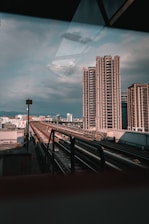 Urban scene with modern high-rise buildings against a cloudy sky. A railway or metro track runs through the center, leading towards the cityscape in the background. The setting conveys a blend of industrial and metropolitan elements.