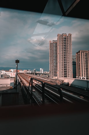 Urban scene with modern high-rise buildings against a cloudy sky. A railway or metro track runs through the center, leading towards the cityscape in the background. The setting conveys a blend of industrial and metropolitan elements.