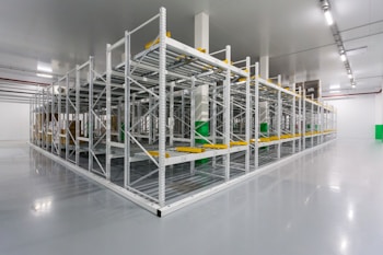 An empty industrial warehouse with metal shelves organized in rows. The polished gray floor reflects the overhead fluorescent lighting. The shelves are sturdy, with yellow guards and identification labels. The environment appears clean and spacious.