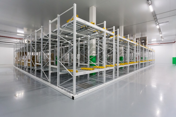 An empty industrial warehouse with metal shelves organized in rows. The polished gray floor reflects the overhead fluorescent lighting. The shelves are sturdy, with yellow guards and identification labels. The environment appears clean and spacious.