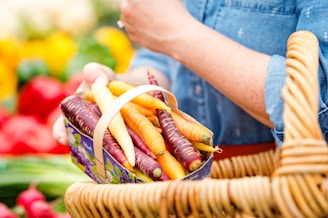 A person holding a small basket filled with multicolored carrots, including purple, orange, and yellow varieties. The person is dressed in a blue denim shirt and is carrying a wicker basket. In the background, there are blurred yellow and red objects, likely other produce at a market.