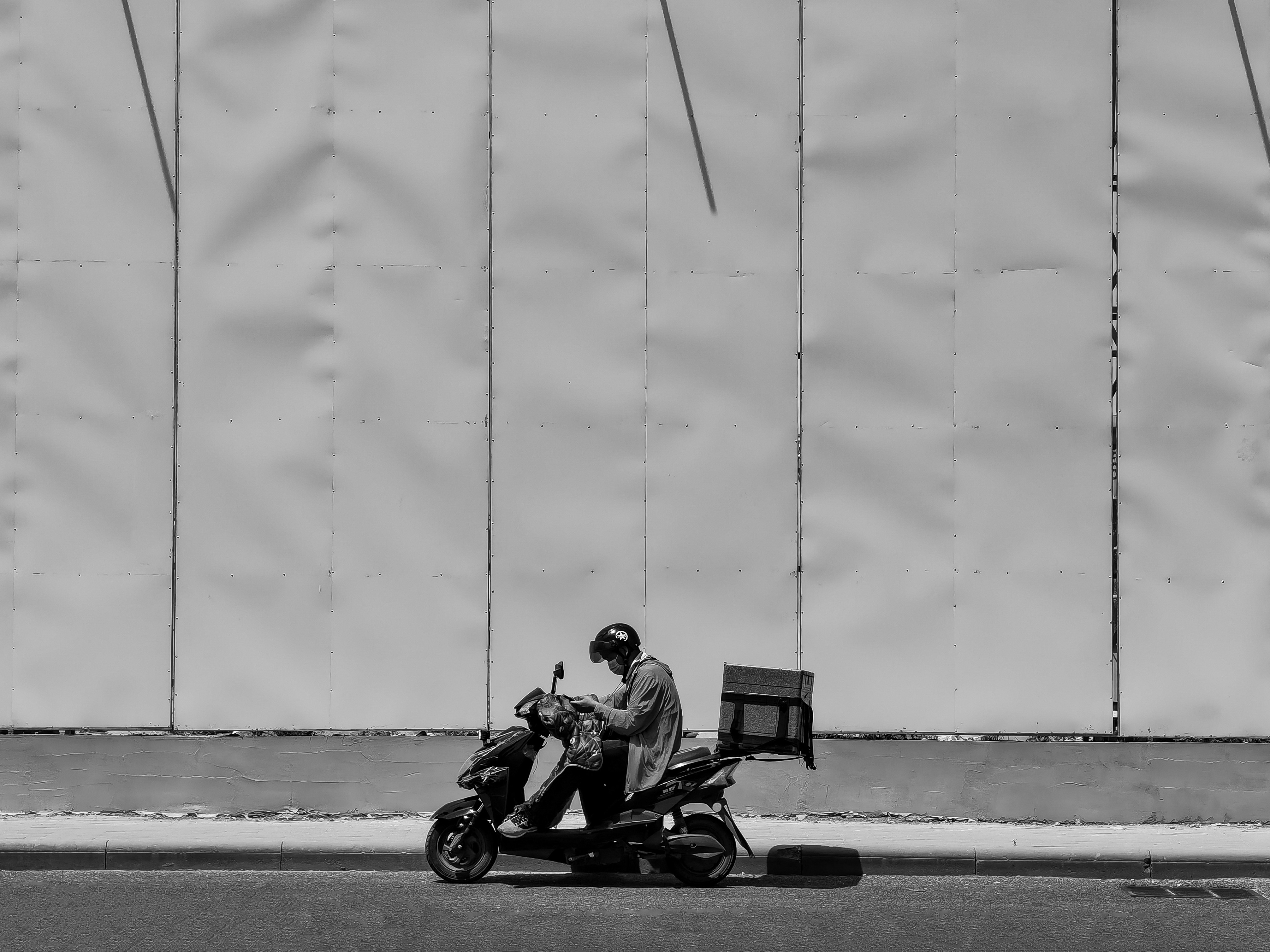A gig worker, identifiable by a delivery bag, sitting dejectedly next to his motorcycle in a long fuel queue.