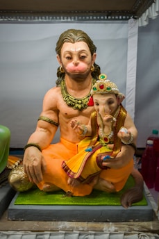 A detailed sculpture of two Hindu deities, with one resembling Hanuman and the other resembling Ganesha. Hanuman is depicted with an orange attire and jewelry. Ganesha, smaller in size, is sitting on Hanuman's lap adorned with a colorful attire and intricate crown with jewels.