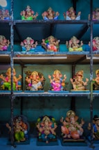 A collection of colorful Ganesha statues displayed on metal shelves. The statues are intricately designed with vibrant garments, adorned with jewelry, and posed with various hand mudras. The background is a textured blue wall, enhancing the vividness of the statues.