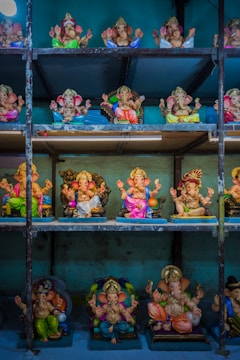A collection of colorful Ganesha statues displayed on metal shelves. The statues are intricately designed with vibrant garments, adorned with jewelry, and posed with various hand mudras. The background is a textured blue wall, enhancing the vividness of the statues.