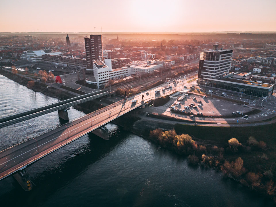 The image presents an aerial view of a cityscape, with a large river flowing through it. The river is bordered by buildings on both sides and stretches across the center of the frame. On one side of the river, there's a bridge that spans its width, connecting two parts of the city. The other side of the river features several tall buildings, including a prominent skyscraper with numerous windows reflecting the sunlight. The sky above is painted in shades of orange and yellow, suggesting either sunrise or sunset.