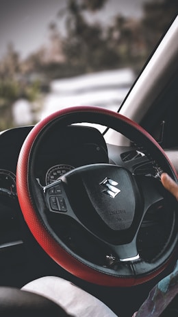 A close-up view of a car's steering wheel with a black and red cover. The logo on the steering wheel indicates the car brand. Behind the wheel, the dashboard and speedometer are visible. The hand of a person is lightly resting on the wheel, and the background outside the car window is slightly blurred, suggesting motion or focus on the foreground.