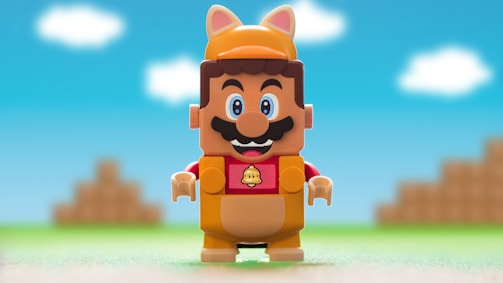 A toy figure resembling a popular video game character dressed in a tanuki suit stands prominently with a cheerful expression. The background features a pixelated, colorful landscape typical of platform games, with a bright blue sky and white clouds.