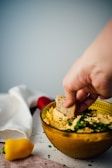 A hand is dipping a piece of bread into a bowl of hummus garnished with chopped herbs. A yellow bell pepper and a white cloth are nearby, adding a casual and inviting atmosphere to the scene.
