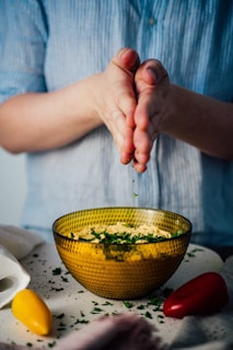 A person wearing a blue shirt sprinkles herbs over a textured yellow bowl filled with a dish. A white cloth and yellow and red vegetables are placed on the surface nearby.