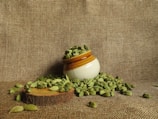 A small ceramic pot filled with green cardamom pods is positioned on a textured brown fabric. Some cardamom pods have spilled around the pot, resting on a wooden slice that serves as a base.