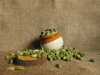 A small ceramic pot filled with green cardamom pods is positioned on a textured brown fabric. Some cardamom pods have spilled around the pot, resting on a wooden slice that serves as a base.