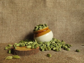 A small ceramic pot filled with green cardamom pods is positioned on a textured brown fabric. Some cardamom pods have spilled around the pot, resting on a wooden slice that serves as a base.