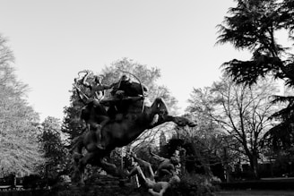 A dramatic black-and-white image capturing a dynamic sculpture of figures in motion, set against a backdrop of leafy trees. The sculpture depicts several human figures and a horse engaged in a vigorous scene, surrounded by trees with intricate branches and leaves.