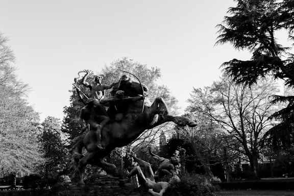 A dramatic black-and-white image capturing a dynamic sculpture of figures in motion, set against a backdrop of leafy trees. The sculpture depicts several human figures and a horse engaged in a vigorous scene, surrounded by trees with intricate branches and leaves.