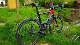 A sleek road bike with a black and red frame is parked on green grass next to a bike rack. The bike features a streamlined design with thin tires. In the background, there are trees, a wooden building, and a pathway.