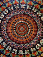 A complex, colorful mandala featuring a circular pattern with intricate, symmetrical designs. The central area is dominated by a concentric arrangement of shapes and colors, creating a kaleidoscopic effect. The outer regions of the mandala are adorned with repeating paisley and floral motifs, showcasing an elaborate and vibrant design.