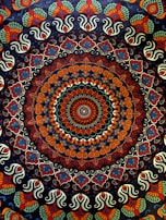 A complex, colorful mandala featuring a circular pattern with intricate, symmetrical designs. The central area is dominated by a concentric arrangement of shapes and colors, creating a kaleidoscopic effect. The outer regions of the mandala are adorned with repeating paisley and floral motifs, showcasing an elaborate and vibrant design.