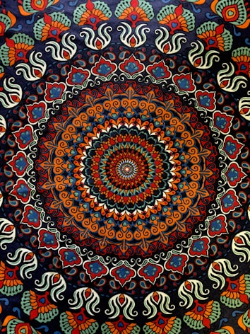 A complex, colorful mandala featuring a circular pattern with intricate, symmetrical designs. The central area is dominated by a concentric arrangement of shapes and colors, creating a kaleidoscopic effect. The outer regions of the mandala are adorned with repeating paisley and floral motifs, showcasing an elaborate and vibrant design.