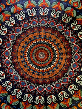 A complex, colorful mandala featuring a circular pattern with intricate, symmetrical designs. The central area is dominated by a concentric arrangement of shapes and colors, creating a kaleidoscopic effect. The outer regions of the mandala are adorned with repeating paisley and floral motifs, showcasing an elaborate and vibrant design.