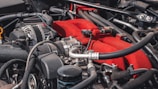 A detailed view of a car engine, featuring various components such as pipes, hoses, and wires. The engine has a prominent red part, likely a manifold cover, standing out against the metallic and black hues of the surrounding components.